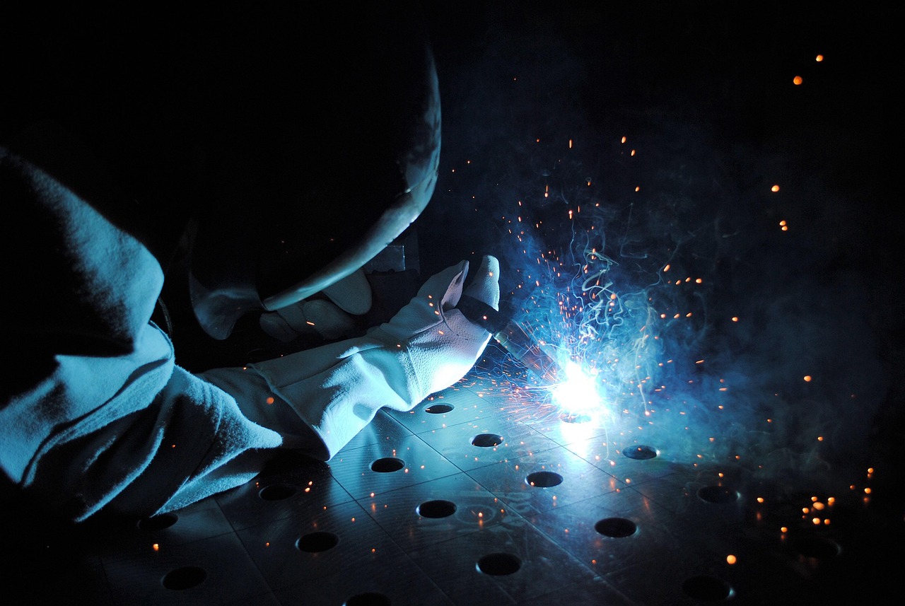 A welder fuses metal outdoors using a welding machine under bright summer sunlight.